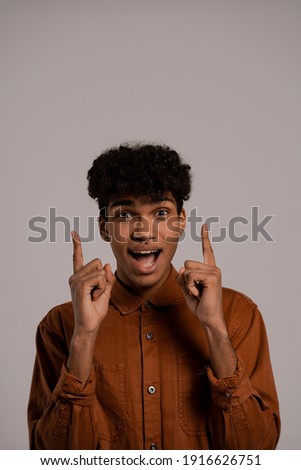Photo of black man shocked and happily surprised about some news, points fingers up. Male wears shirt, isolated grey color background