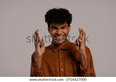 Photo of black man crosses fingers and looks worried about something. Male wears shirt, isolated grey color background