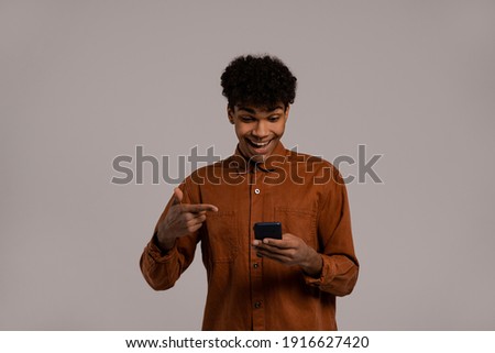Photo of black man holds smartphones and points at it, looks very amazed. Male wears shirt, isolated grey color background