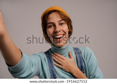 Photo of cute female hipster takes a selfie and smile with nice teeth. White woman wears denim overall and hat isolated grey color background