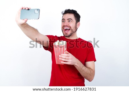 Young handsome man in red T-shirt against white background eating popcorn taking a selfie to post it on social media or having a video call with friends.