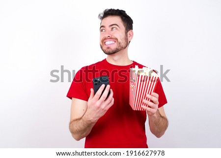 Young handsome man in red T-shirt against white background eating popcorn holding in hands showing new cell