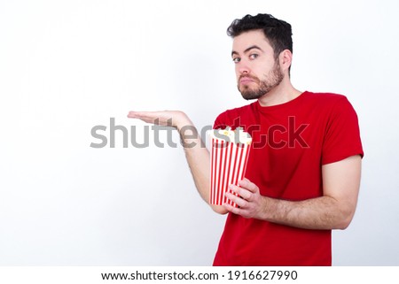 Young handsome man in red T-shirt against white background eating popcorn pointing aside with both hands showing something strange and saying: I don't know what is this. Advertisement concept.