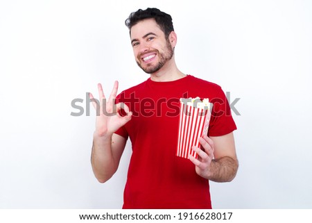 Young handsome man in red T-shirt against white background eating popcorn showing both hands with fingers in OK sign. Approval or recommending concept