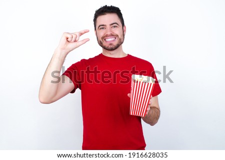 Young handsome man in red T-shirt against white background eating popcorn smiling and confident gesturing with hand doing small size sign with fingers looking and the camera. Measure concept