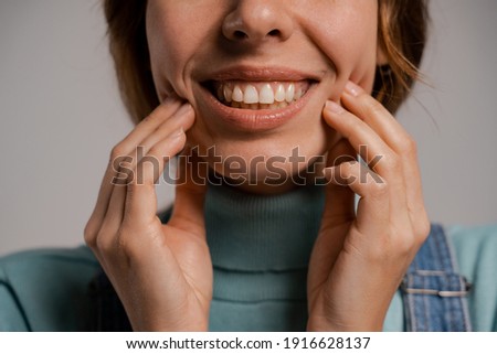 Photo of cute female hipster holds face and looks like she has nice teeth. White woman wears denim overall and hat isolated grey color background
