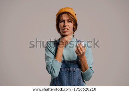 Photo of cute female hipster has sore throat and looks like got sick. White woman wears denim overall and hat isolated grey color background