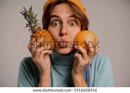 Photo of cute female hipster play with pineapple and orange, looks healthy. White woman wears denim overall and hat isolated grey color background