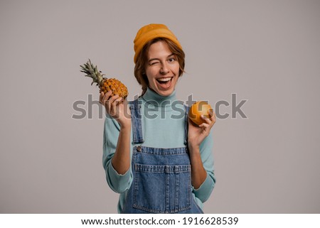 Photo of cute female hipster plays with pineapple and orange, looks healthy. White woman wears denim overall and hat isolated grey color background