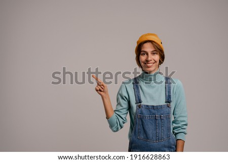 Photo of cute female hipster smiles and points at left side. White woman wears denim overall and hat isolated grey color background