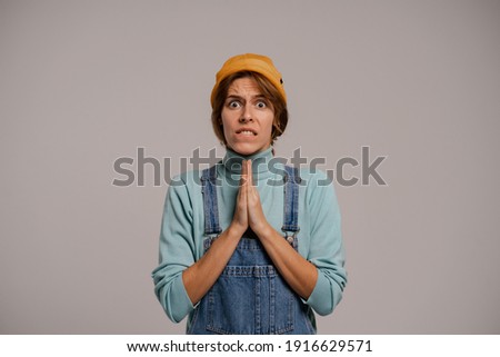Photo of cute female hipster prays and waits for something so much. White woman wears denim overall and hat isolated grey color background