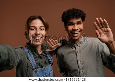 Photo of cute couple farmers takes a selfie with nice smile. Woman wears denim overalls, man wears t-shirt, isolated brown color background