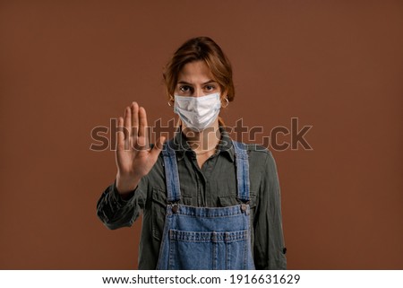 Photo of cute female farmer with face mask shows lockdown and quarantine time. Wears denim overalls, isolated brown color background.