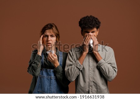 Photo of cute couple farmers, both blow their nose and got sick. Female wears denim overalls, male wears t-shirt isolated brown color background