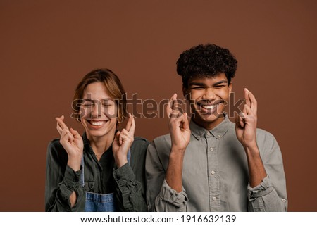 Photo of cute couple farmers cross their fingers to pray for something. Female wears denim overalls, male wears t-shirt, isolated brown color background