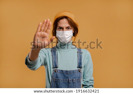 Photo of cute female farmer with face mask for quarantine time show to keep distance. Wears denim overalls and hat, isolated brown color background.