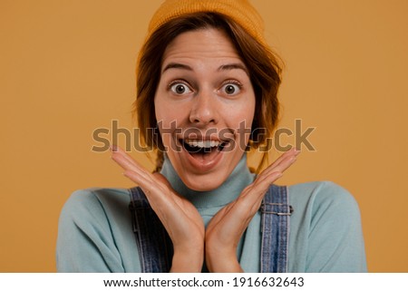 Close up photo of cute female farmer surprised about something. Wears denim overalls and hat, isolated brown color background