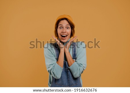Photo of cute female farmer surprised about something. Wears denim overalls and hat, isolated brown color background