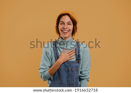 Photo of cute female farmer surprised and smiles about something, looks happy. Wears denim overalls and hat, isolated brown color background