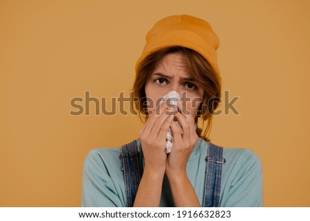 Photo of cute female farmer has got running nose and looks sick. Wears denim overalls and hat, isolated brown color background.
