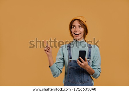 Photo of cute female farmer holds smartphone points at left side of picture. Wears denim overalls and hat, isolated brown color background
