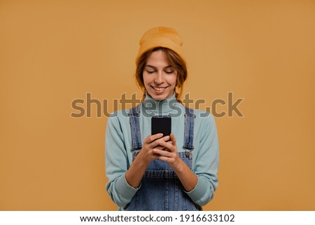 Photo of cute female farmer holds smartphone and seems happy. Wears denim overalls and hat, isolated brown color background