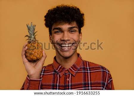 Photo of cute African male farmer holds pineapple and slime. Man wears plaid shirt, isolated brown color background
