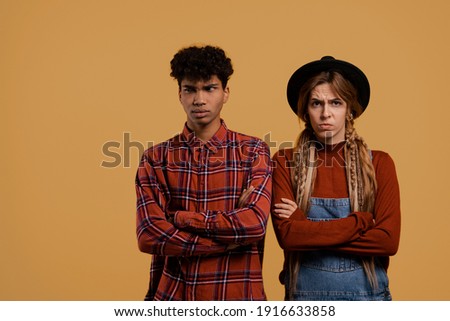 Photo of cute couple farmers argue about something and don't talk to each other. Black man wears plaid shirt, white woman wears denim overall and hat isolated brown color background
