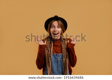 Photo of cute female farmer looks surprised and happy. White woman wears denim overall and hat isolated brown color background