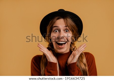 Photo of cute female farmer smiles, looks funny and surprised. White woman wears denim overall and hat isolated brown color background