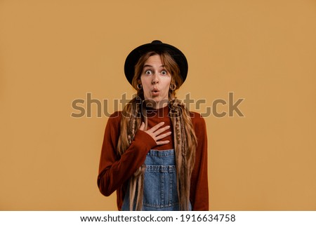 Photo of cute female farmer looks shocked. White woman wears denim overall and hat isolated brown color background