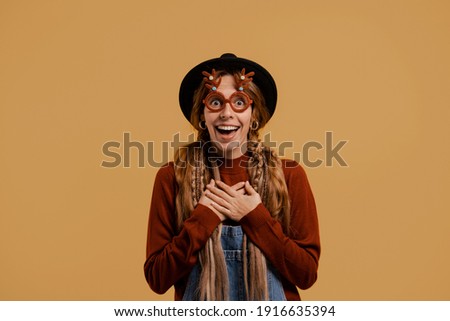 Photo of attractive female farmer stands, wears funny glasses and looks very surprised. White woman wears denim overall and hat isolated brown color background