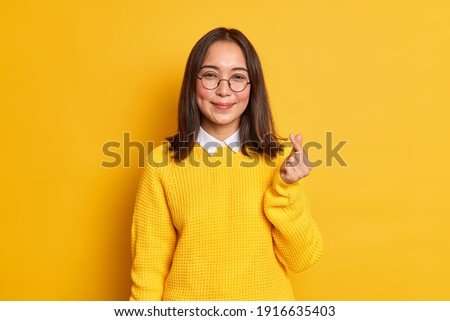 Lovely pleased Asian woman makes korean like gesture smiles gently expresses love to someone wears round transparent glasses and knitted sweater isolated over yellow background. Body language
