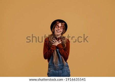 Photo of attractive female farmer looks pleasant and happy, wears funny glasses. White woman wears denim overall and hat isolated brown color background