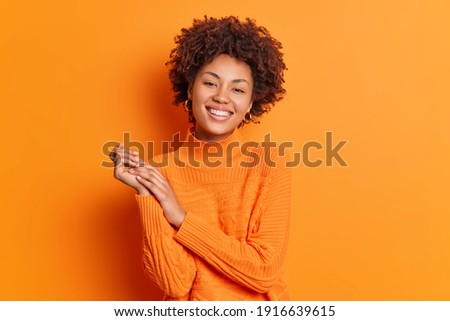 Pretty curly haired young woman smiles gently keeps hands together focused directly at camera has pleasant talk with best friend dressed in casual sweater isolated over vivid orange background