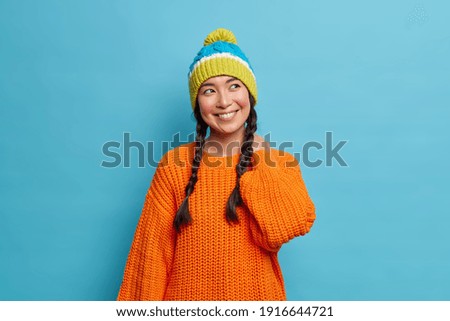 Dreamy pretty Asian girl has two combed pigtails looks happily aside thinks about something pleasant wears orange knitted sweater and warm hat isolated over blue background. Winter style concept