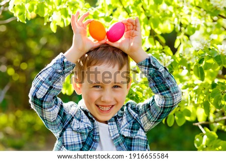 A boy on an Easter egg hunt in a blooming spring garden. He holds the decorated eggs to his head like a rabbit's ears.