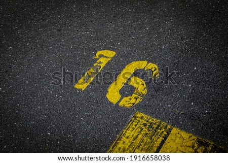Grey asphalt road, yellow painted number sixteen on grey street, yellow lines like a corner on the road, space for text 
