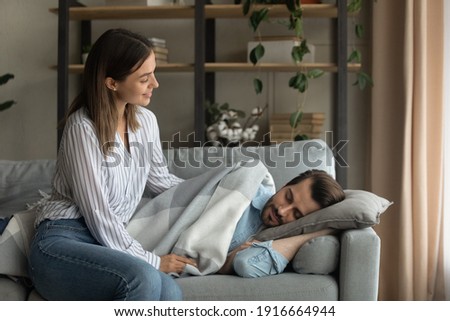 Love in details. Attentive millennial wife carefully covering tired husband sleeping on couch with warm soft plaid blanket. Affectionate young female take care of fatigued spouse resting on cozy sofa Royalty-Free Stock Photo #1916664944