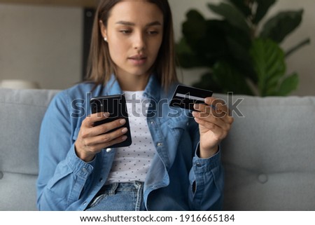 Unsuspecting young lady dubious web shop client making purchase at internet giving personal data to fraudulent scam website using cell. Soft focus on woman hands with modern mobile phone and bank card Royalty-Free Stock Photo #1916665184