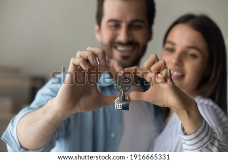 Love lives here. Happy caring millennial family couple posing at new home house cuddling making heart shape of fingers with bunch of keys inside. Focus on joined hands of spouses holding key. Close up Royalty-Free Stock Photo #1916665331