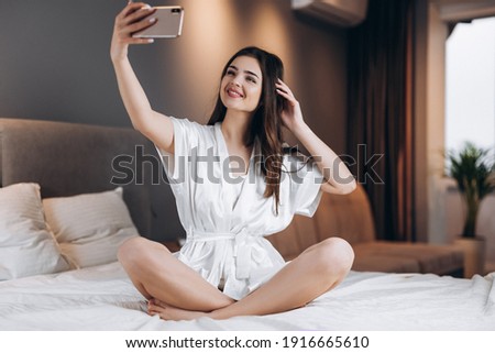 Young beautiful woman in white silk pajamas makes a selfie on the phone. A female model with a phone in her hands sits on the bed. morning or evening in the bedroom with a female model