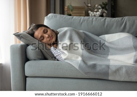 Sweet dreams. Serene millennial female lie on soft cozy couch in comfortable pose hidden under warm woolen blanket having tight sleep. Satisfied young lady nap on sofa at home enjoy good rest relax Royalty-Free Stock Photo #1916666012