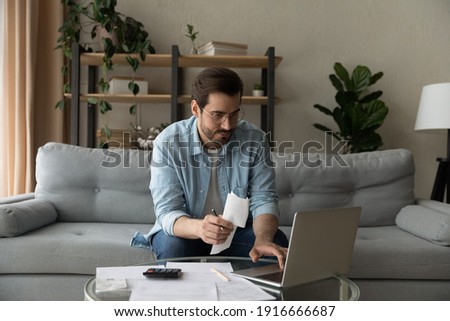 Serious millennial male in glasses focused on paying utility bills taxes rental charges online using laptop. Young man working from home office sit on couch hold paper invoice check information online Royalty-Free Stock Photo #1916666687