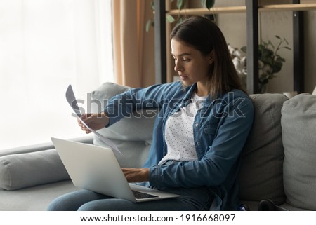 Serious millennial woman sit on couch hold laptop work with electronic document and paper hardcopy of loan insurance contract. Focused young female read official letter check actual information online Royalty-Free Stock Photo #1916668097