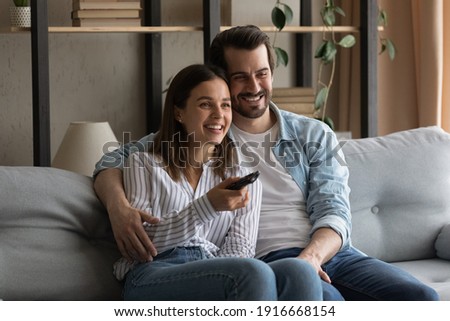 Common interests. Bonding millennial family couple hugging on sofa at living room having pleasure watching movie on tv. Friendly loving young spouses enjoy video film soccer match on weekend evening Royalty-Free Stock Photo #1916668154