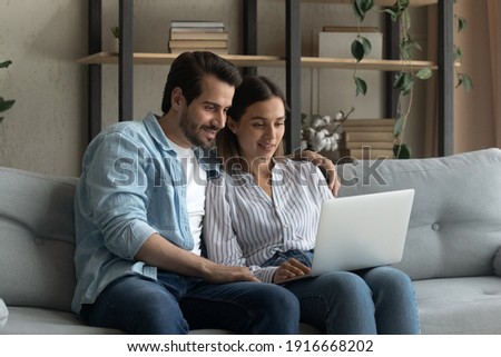 Happy young husband wife spend evening at home relax on couch hold laptop on knees watch video read news having fun play online game. Friendly loving family couple engaged in virtual shopping using pc