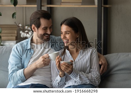 Happy married couple relax on cozy sofa at home cuddle discuss good news drink aromatic tea strong coffee. Smiling young man woman in love meet morning together sit on couch hold cups with hot drinks Royalty-Free Stock Photo #1916668298