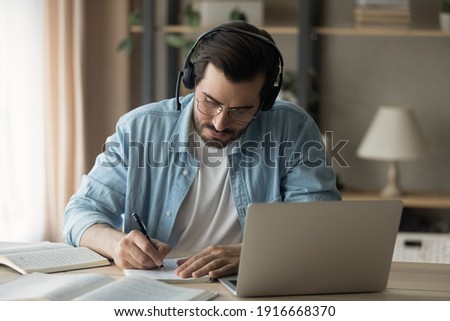 Young guy in modern headset study from home using laptop take test exam make list of answers on questions proposed by teacher online. Millennial man interpreter focused on remote work make translation