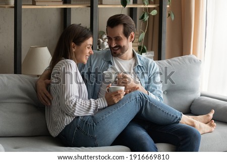 Nothing is better. Affectionate lovers spouses cuddle on couch at living room have pleasure joke laugh drink tea coffee. Happy young husband wife renters tenants of new flat enjoy cozy evening at home Royalty-Free Stock Photo #1916669810